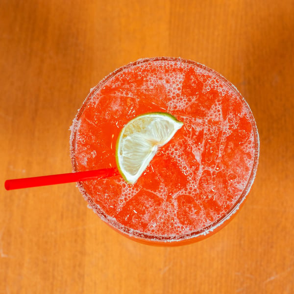 Overhead view of a vibrant red margarita with a lime wedge and red straw, on an orange surface.