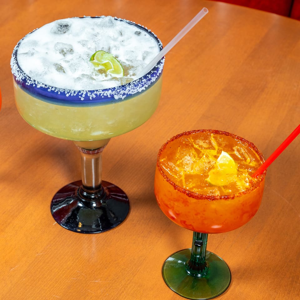 Two margaritas, one classic lime and one orange with chili rim, on a wooden table.