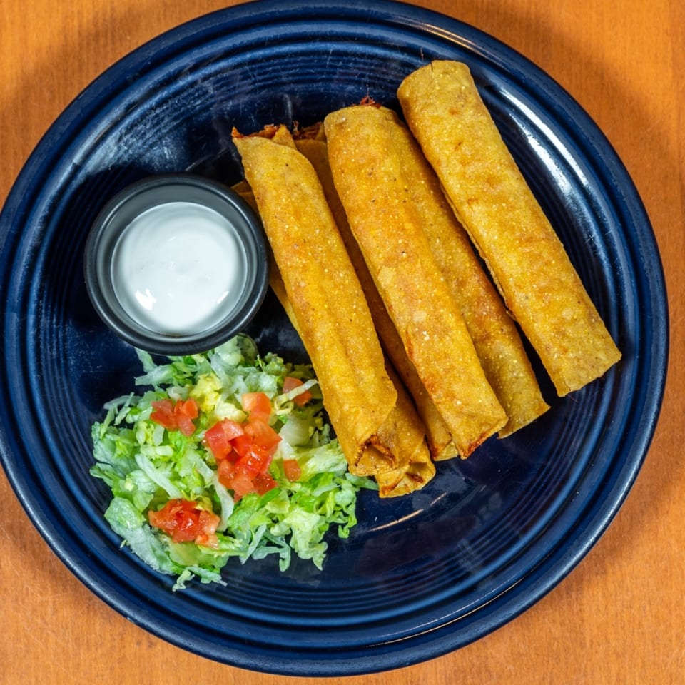 Golden-fried flautas with shredded lettuce, diced tomatoes, and a side of sour cream on a blue plate.