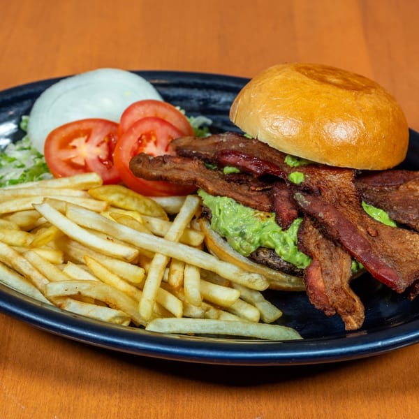 A Mi Cocina bacon guacamole burger with fries, lettuce, tomato, and onion on a dark plate.
