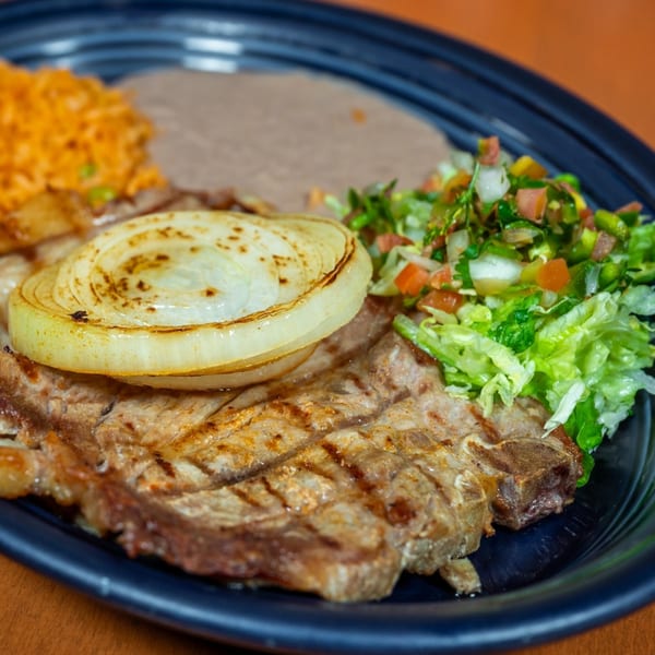 Grilled steak with a large onion slice, rice, refried beans, and a side salad on a black plate.